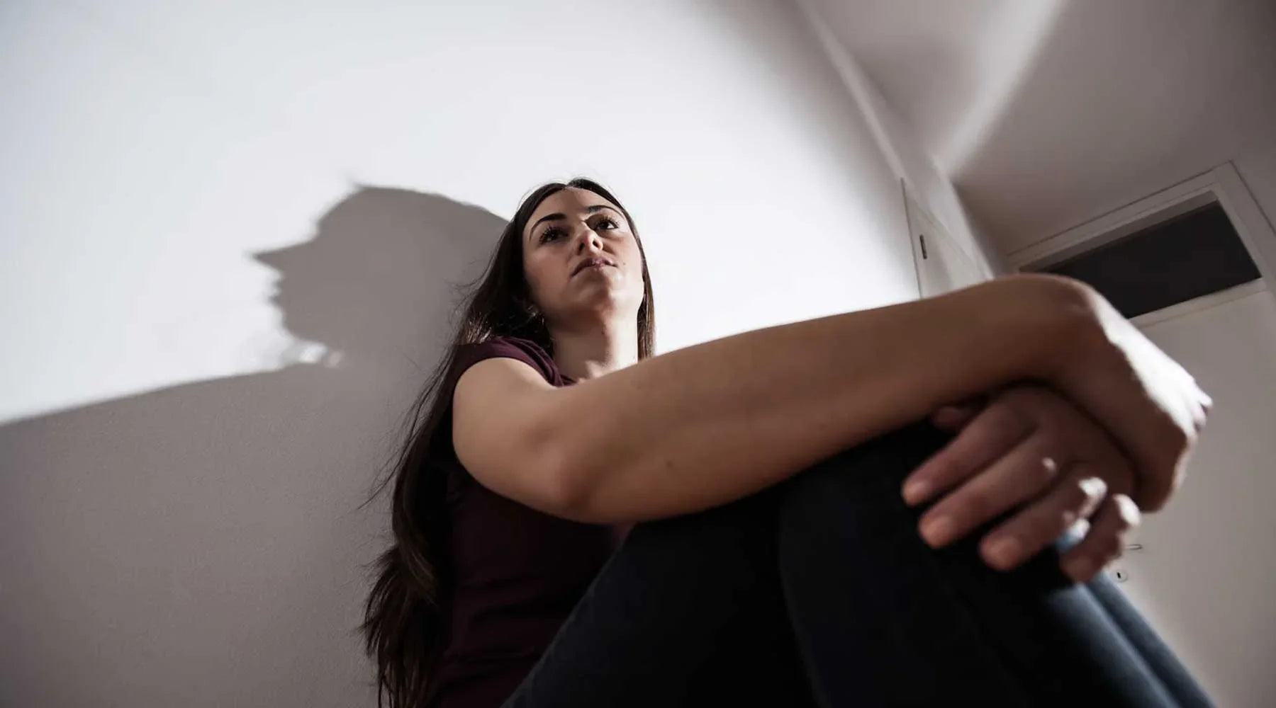 Mujer sentada en el suelo interior, con expresión pensativa, relacionada al bienestar y ansiedad.